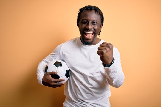 Young african american player man playing soccer holding football ball over yellow background screaming proud and celebrating victory and success very excited, cheering emotion