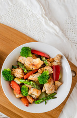 Zenith view of a bowl with fried breast chicken, asparagus, broccoli, mushrooms, red peppers and potatoes on a white table. 