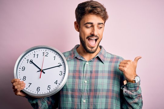 Young man with beard doing countdown using big clock over isolated pink background pointing and showing with thumb up to the side with happy face smiling