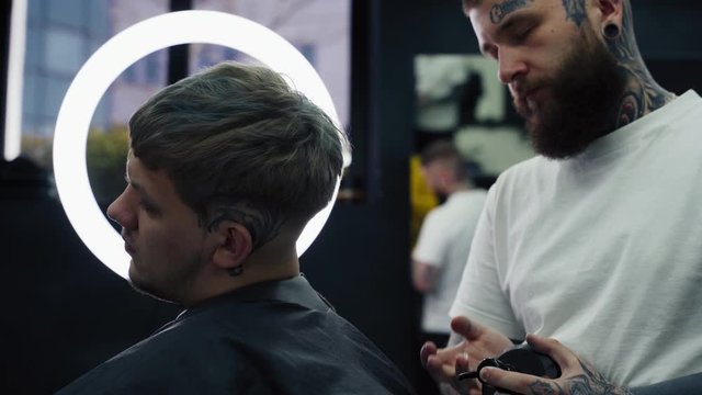 Barber makes hair styling with hair spray after haircut at the barber shop. Young handsome Caucasian man getting a haircut in a modern hairsalon. Handheld shot. 4K.