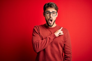 Young handsome man with beard wearing glasses and sweater standing over red background Surprised pointing with finger to the side, open mouth amazed expression.