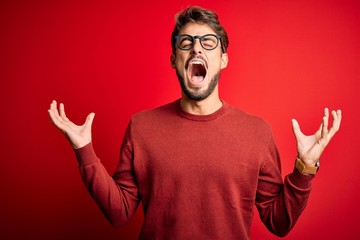 Fototapeta premium Young handsome man with beard wearing glasses and sweater standing over red background crazy and mad shouting and yelling with aggressive expression and arms raised. Frustration concept.