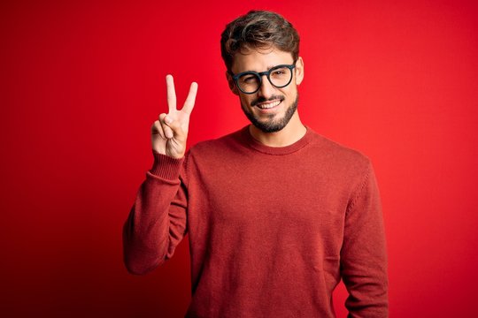 Young handsome man with beard wearing glasses and sweater standing over red background smiling looking to the camera showing fingers doing victory sign. Number two.
