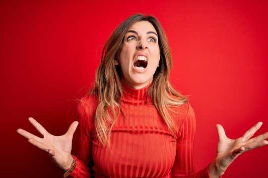 Young beautiful brunette woman wearing casual turtleneck sweater over red background crazy and mad shouting and yelling with aggressive expression and arms raised. Frustration concept.