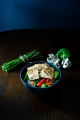 Bol con salmón salvaje, arroz, esparragos trigueros, brócoli, pimiento rojo y champiñones sobre mesa marrón. Dark photography. Fondo oscuro. 