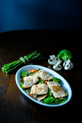 Fuente con salmón salvaje, arroz, esparragos trigueros, brócoli, pimiento rojo y champiñones sobre mesa marrón. Dark photography. Fondo oscuro. 