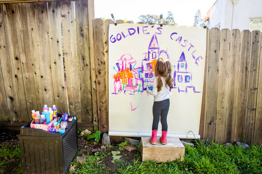 A young girl painting a picture of a castle outside on the side of a fence.
