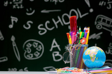 School background with stationery accessories. Books, globe, pencils and various office supplies lying on the desk on a green blackboard background
