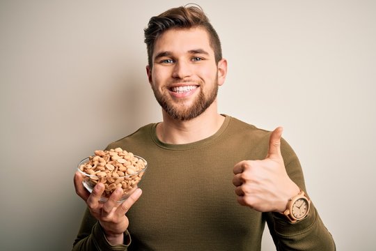 Young blond man with beard and blue eyes holding bowl with healthy salty peanuts happy with big smile doing ok sign, thumb up with fingers, excellent sign
