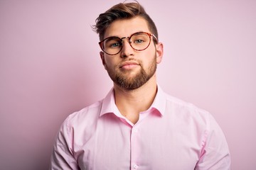 Obraz premium Young handsome blond man with beard and blue eyes wearing pink shirt and glasses Relaxed with serious expression on face. Simple and natural looking at the camera.