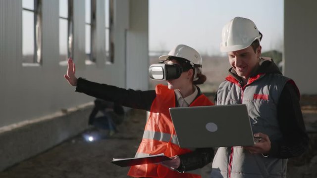 modern technology, woman engineer in a protective helmet and viar glasses looks at the mock-up of a new hangar together with a male colleague in a helmet and a laptop in his hands