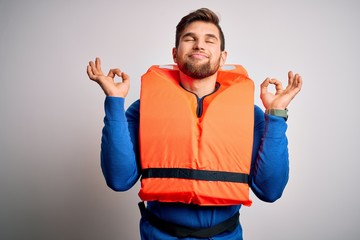 Young blond tourist man with beard and blue eyes wearing lifejacket over white background relax and smiling with eyes closed doing meditation gesture with fingers. Yoga concept.