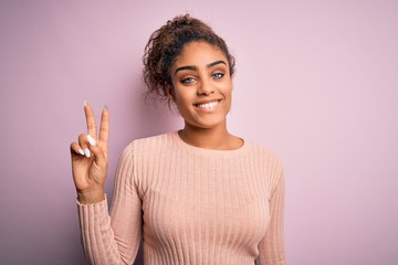 Young beautiful african american girl wearing casual sweater standing over pink background smiling looking to the camera showing fingers doing victory sign. Number two.