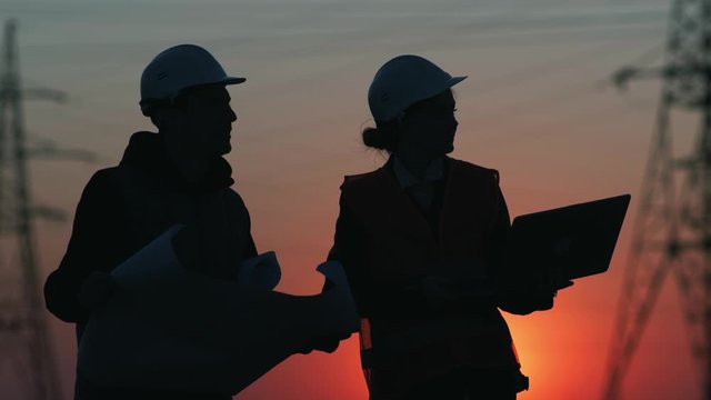 female and male engineers of a high-voltage power station with a laptop and drawings discuss a plan for supplying electricity to the city at sunset, silhouette
