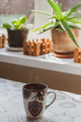 A steaming cup of hot tea or coffee in the early morning on a table near the window. Plants on the windowsill