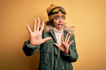 Young african american afro skier girl wearing snow sportswear and ski goggles afraid and terrified with fear expression stop gesture with hands, shouting in shock. Panic concept.