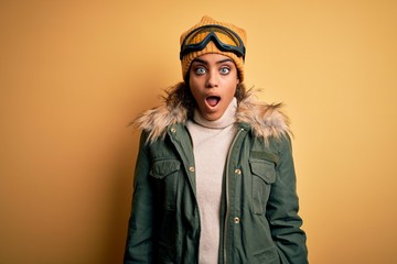 Young african american afro skier girl wearing snow sportswear and ski goggles afraid and shocked with surprise and amazed expression, fear and excited face.