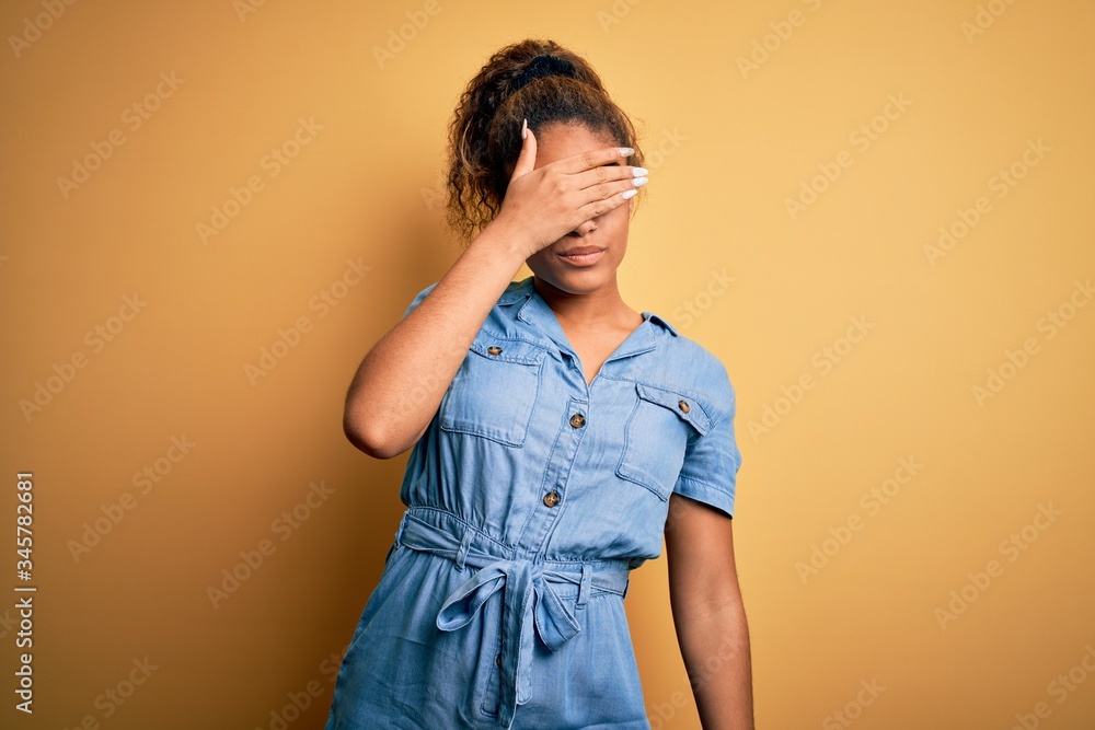 Sticker Young beautiful african american girl wearing denim dress standing over yellow background covering eyes with hand, looking serious and sad. Sightless, hiding and rejection concept