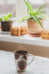 A steaming cup of hot tea or coffee in the early morning on a table near the window. Plants on the windowsill