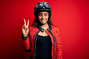 Young african american motorcyclist girl wearing moto helmet and glasses over red background smiling with happy face winking at the camera doing victory sign. Number two.