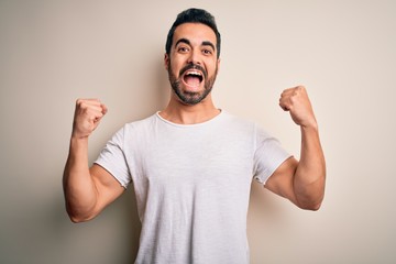 Naklejka premium Young handsome man with beard wearing casual t-shirt standing over white background celebrating surprised and amazed for success with arms raised and open eyes. Winner concept.