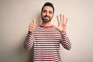 Fototapeta premium Young handsome man with beard wearing casual striped t-shirt standing over white background showing and pointing up with fingers number six while smiling confident and happy.