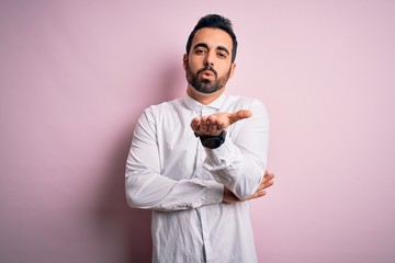 Young handsome man with beard wearing casual shirt standing over pink background looking at the camera blowing a kiss with hand on air being lovely and sexy. Love expression.