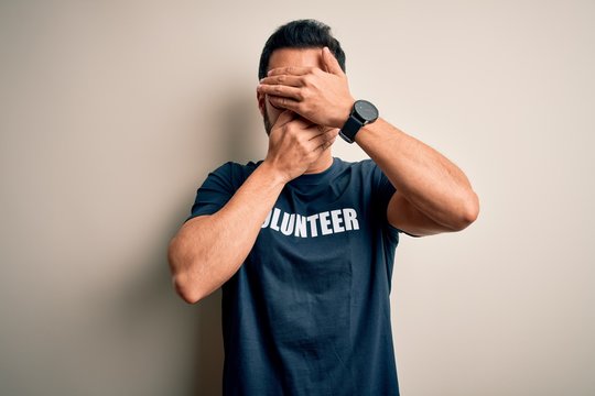 Handsome man with beard wearing t-shirt with volunteer message over white background Covering eyes and mouth with hands, surprised and shocked. Hiding emotion