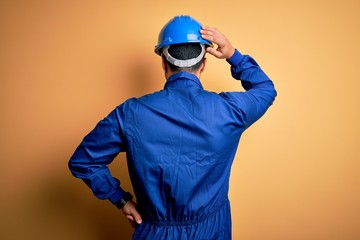 Mechanic man with beard wearing blue uniform and safety helmet over yellow background Backwards thinking about doubt with hand on head