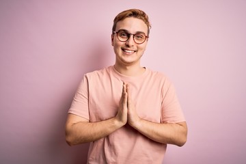 Young handsome redhead man wearing casual t-shirt standing over isolated pink background praying with hands together asking for forgiveness smiling confident.