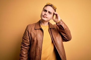Young handsome redhead man wearing casual leather jacket over isolated yellow background confuse...