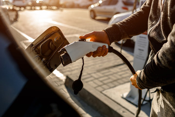 Man Holding Power Charging Cable For Electric Car In Outdoor Car Park. And he s going to connect the car to the charging station in the parking lot near the shopping center