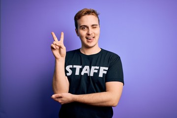 Fototapeta premium Young handsome redhead worker man wearing staff t-shirt uniform over purple background smiling with happy face winking at the camera doing victory sign with fingers. Number two.