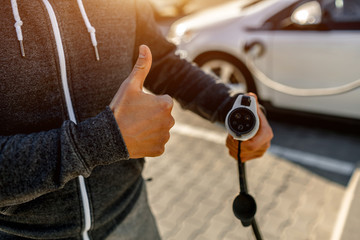 Man Holding Power Charging Cable For Electric Car In Outdoor Car Park. The driver a thumb up