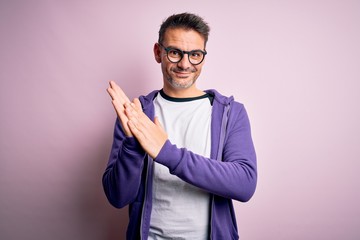 Young handsome man wearing purple sweatshirt and glasses standing over pink background clapping and applauding happy and joyful, smiling proud hands together