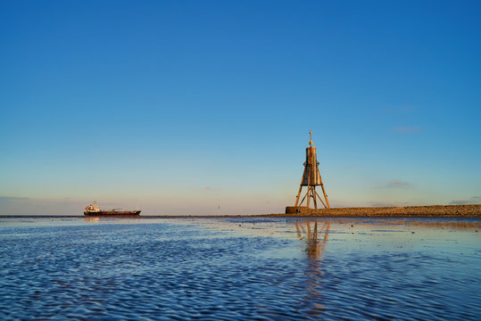 the historic beacon "Kugelbake" in Cuxhaven (Germany) seen from the mud flats of the Wadden Sea in beautiful late afternoon sunlight against blue sky, a medium size freight ship is heading to the Elbe