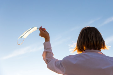 Happy woman takes off medical protective mask holds it on her finger on blue sky background, enjoys...