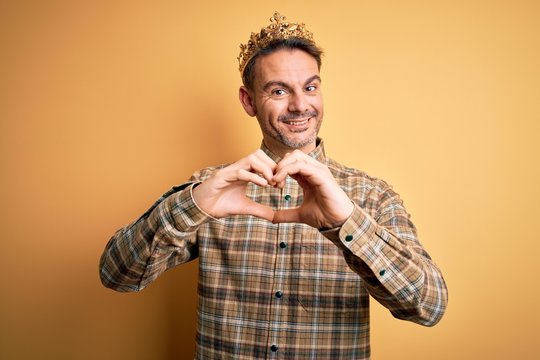 Young handsome man wearing golden crown of king over isolated yellow background smiling in love showing heart symbol and shape with hands. Romantic concept.