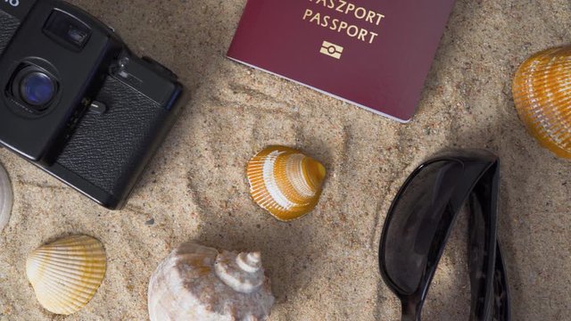 Passport, vintage film camera, sunglasses and seashells on golden beach sand. Summer travel abstract concept. Panning shot.
