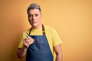 Young handsome blonde hairdresser man wearing apron holding scissors standing over isolated yellow background