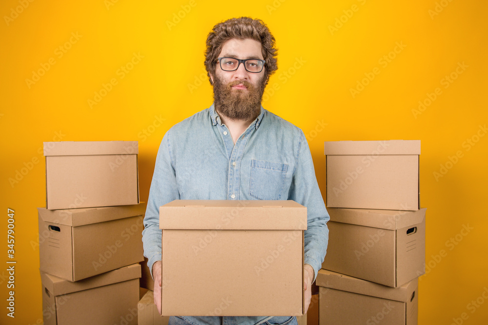 Wall mural Bearded man delivery man holding a corton box in his hands, standing among parcels on a yellow background