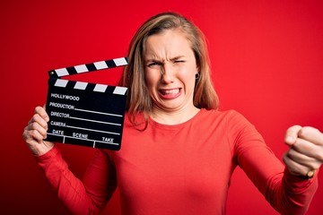 Young beautiful blonde clapper woman holding clapboard over isolated red background annoyed and...