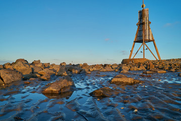 the historic beacon "Kugelbake" in Cuxhaven (Germany) seen in frog perspective from the mud flats of the Wadden Sea next to the groyne in beautiful late afternoon sunlight against blue sky