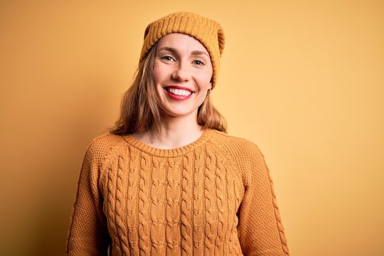 Young beautiful blonde woman wearing casual sweater and wool cap over yellow background with a happy and cool smile on face. Lucky person.