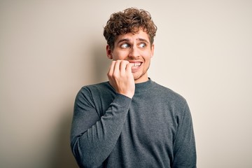 Young blond handsome man with curly hair wearing casual sweater over white background looking stressed and nervous with hands on mouth biting nails. Anxiety problem.