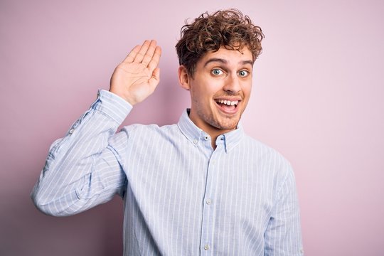 Young blond handsome man with curly hair wearing striped shirt over white background Waiving saying hello happy and smiling, friendly welcome gesture