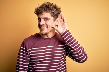 Young blond handsome man with curly hair wearing casual striped sweater smiling with hand over ear listening an hearing to rumor or gossip. Deafness concept.