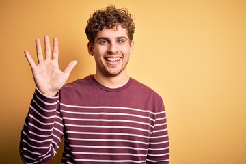 Young blond handsome man with curly hair wearing casual striped sweater showing and pointing up with fingers number five while smiling confident and happy.