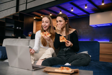 Slow motion of surprised appealing 25s stylish female friends which sitting on the comfortable sofa and enjoying delicious pizza during browsing amusing pictures on computer