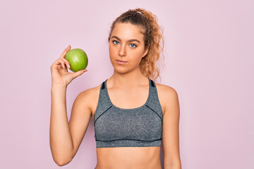 Young beautiful blonde sporty woman with blue eyes holding healthy green apple fruit with a confident expression on smart face thinking serious